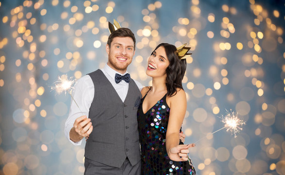 celebration, fun and holidays concept - happy couple with crowns and sparklers at party over festive lights background