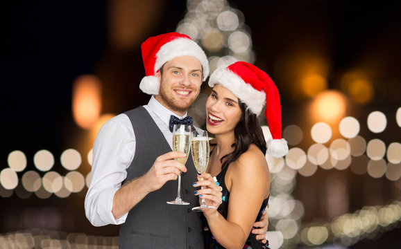 Celebration, People And Holidays Concept - Happy Couple In Santa Hats With Glasses Of Non Alcoholic Champagne Over Christmas Tree Lights Background