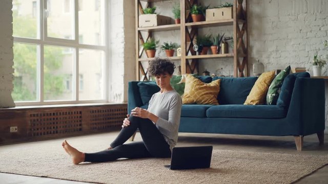 Pretty young lady is learning yoga from online lessons sitting on floor looking at laptop screen and practising body twists. Internet, sports and hobby concept.