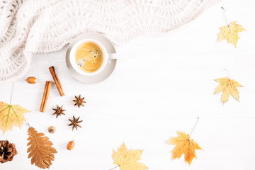 Autumn or winter cozy composition. Cup of coffee, warm knitted plaid, cinnamon sticks, anise star and dry leaves on a white background wooden table. Flat lay, top view, copy space 