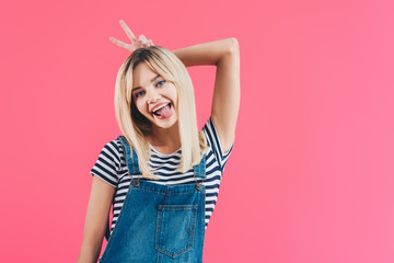 smiling beautiful girl in denim overall sticking tongue out and showing bunny ears isolated on pink