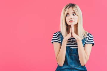 sad beautiful girl in denim overall looking away with hands in namaste gesture isolated on pink