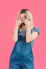 beautiful shocked girl in denim overall looking at camera through fingers isolated on pink