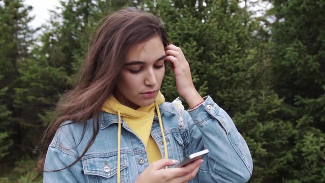 Young teen coed using touch screen smartphone while in nature. Hipster using social media and smiling while reading mobile. Beautiful woman in denim jacket in spruce forest