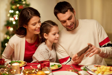 holidays, family and technology concept - happy mother, father and little daughter with smartphone having christmas dinner at home