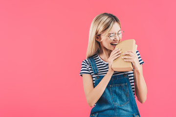 portrait of young student in eyeglasses biting books isolated on pink