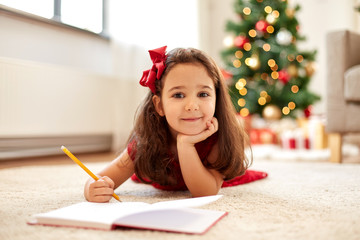 christmas, holidays and childhood concept - smiling girl writing wish list or letter to santa at...