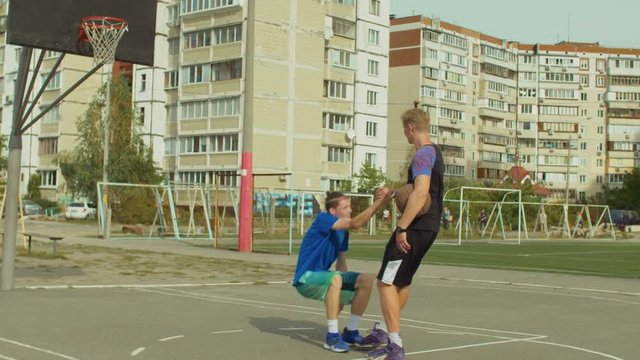 Streetball player with ball helping fallen opponent to get up from basketball court after committed foul while playing one on one streetball game. Man extending hand to lift opponent fallen on ground.