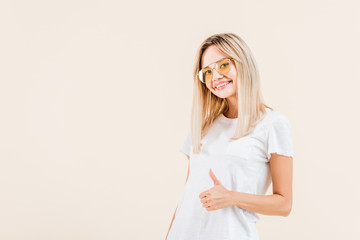 happy young woman in sunglasses showing thumb up and smiling at camera isolated on beige