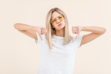 unsatisfied young woman in sunglasses showing thumbs down and looking at camera isolated on beige