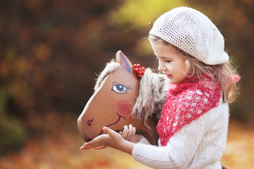 Cute little girl feeds a wooden toy horse in the autumn garden. © fadzeyeva