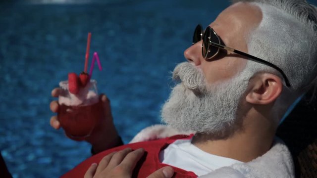 Happiness and satisfaction at the tropical new year. Crazy cheerful traveler chilling on sunbathing bad in santa costume, tanning on the sun, drinking alcohol with straw near with hotel swimming pool