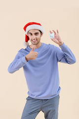 Fototapeta na wymiar happy young man in santa hat holding condom and showing thumb up isolated on beige