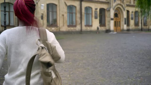 Young african student late for classes and running to university, woman with pink dreadlocks holding backpack