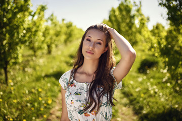Fototapeta premium Potrait of happy brunette who is touching her hair and standing in a fruit orchard.
