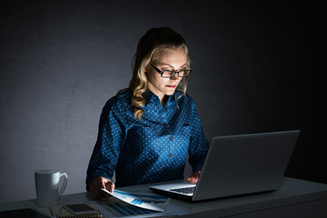 Attractive blonde working on laptop in dark office. Mixed media