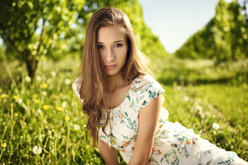 Naklejka na ściany i meble Portrait of a long-haired girl in a summer dress, with green orchard in the background.