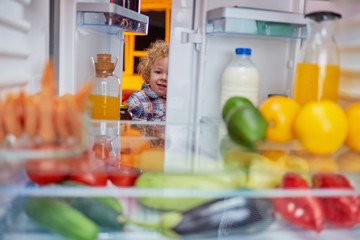 Toddler peeking in the fridge full of groceries and looking something to eat. Picture taken from the inside of fridge.