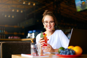 Smiling mixed race woman with cocktail in hand works with laptop. Businesswoman in glasses drinks...