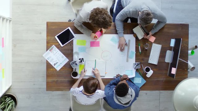 Top view of group of four multiethnic business people sitting at desk in office and using sticky notes to create mind map on large sheet of paper
