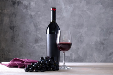 Vintage bottle of red wine with blank matte black label, bunch of grapes on wooden table, concrete wall background. Expensive bottle of cabernet sauvignon concept. Copy space, top view, flat lay.