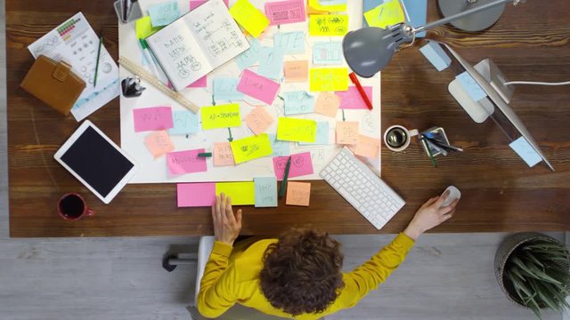 Top view of concentrated young curly haired woman sitting at desk in modern office and writing messages on sticky notes