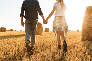 Cropped photo of guy and girl walking hand by hand through golden field with bunch of haystacks, during sunny day