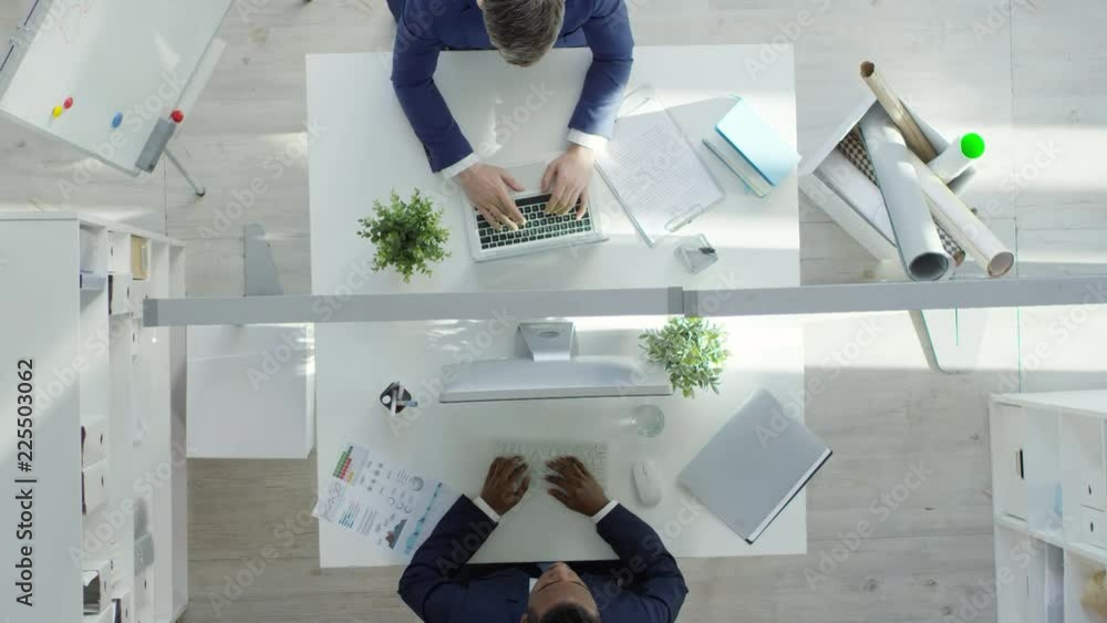 Poster top view of two male employees of different ethnicities using computers while sitting opposite each 