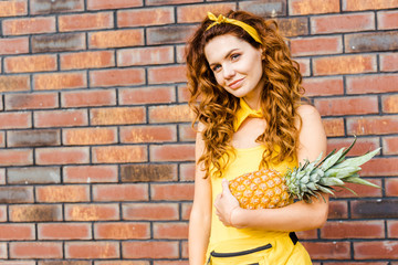 attractive young woman in yellow clothes holding pineapple in front of brick wall and looking at camera