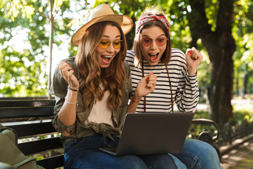Excited happy ladies friends outdoors sitting using laptop computer.