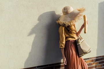fashionable woman in straw hat with baguette standing in front of white wall