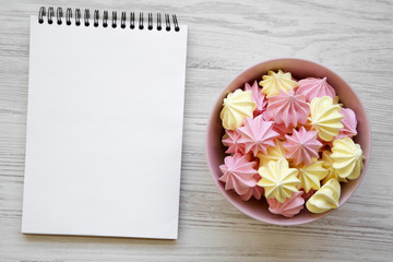 Mini meringues in a pink bowl and blank notebook over white wooden background, top view. Flat lay, overhead, from above.