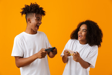 Excited young cute african couple isolated over yellow background play games by mobile phones.