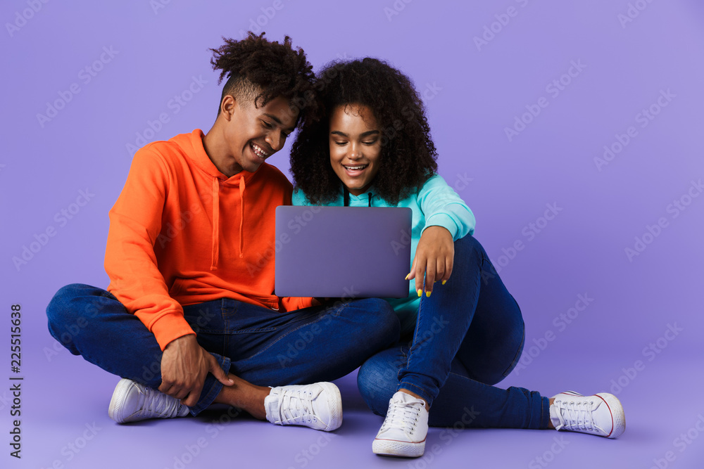 Sticker Cute african couple sitting isolated over violet background using laptop computer.