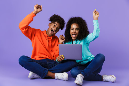 Happy young cute african couple sitting isolated over violet background using laptop computer make winner gesture.