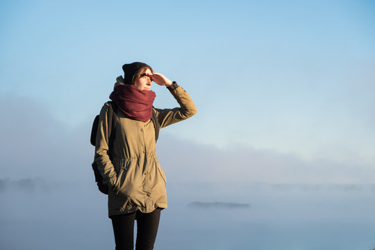 Woman stands in morning sunlight in front of beautiful nature scenery covered in fog. Female hiker enjoying rising sun in misty natural background