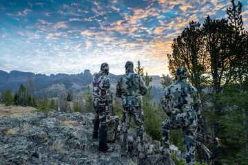  Three adult male hunter friends, unrecognizable,  stand on a mountain ridge looking for elk to hunt during bow archery season. Wearing camouflage © MelissaMN