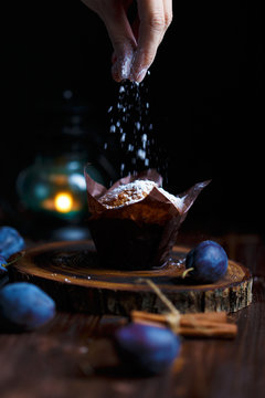 Female hands sprinkled with blueberry muffin with powdered sugar on a dark wooden background with plums.