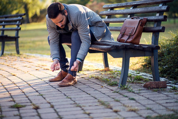 businessman tying shoes while sitting on a bench in a park