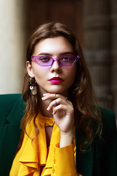 Outdoor close up portrait of young beautiful fashionable woman wearing trendy violet color sunglasses, earrings, stylish clothes. Model looking at camera. Female fashion concept. 