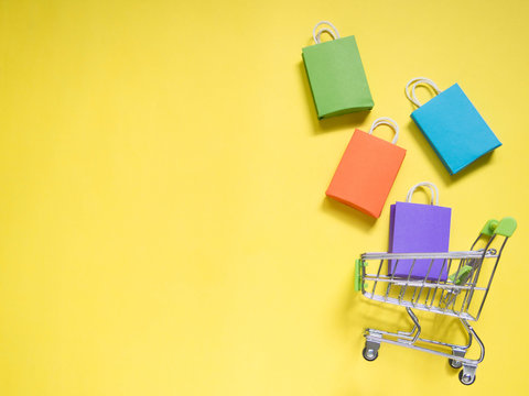Trolley cart and colorful paper shopping bags on yellow background. Creative idea for shopping online, summer sale, supermarket, discount promotion, Cyber Monday and Black Friday concept. Copy space .