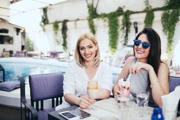 Two young and beautiful woman enjoying in private cocktail house party. They smiling and looking at camera. Swimming pool in background.