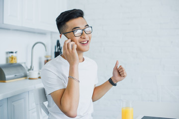 happy young asian man talking by smartphone while sitting in kitchen