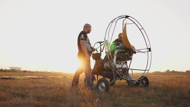 Motor paraglider stands in a field at sunset with a wooden propeller, two pilots warm up the engine before the flight. A test run of all systems