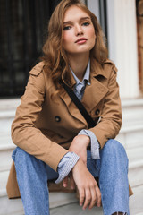 Nice girl in trench coat and jeans dreamily looking in camera sitting on little stairs while...