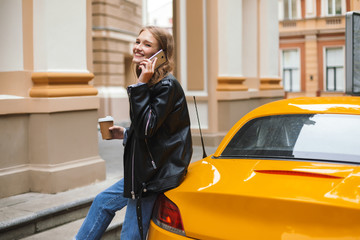 Pretty cheerful girl in leather jacket with cup of coffee to go leaning on yellow sport car talking on cellphone while joyfully looking in camera on city street