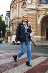 Young cheerful woman in leather jacket and jeans happily looking in camera holding cup of coffee to go in hand while walking on cozy city street