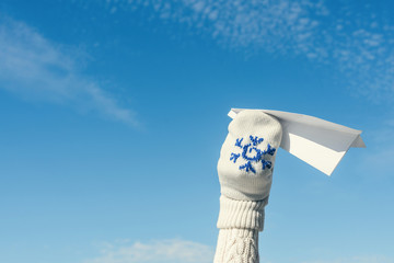 A girl in the winter knitted mittens is launching a paper airplane on the blue sky background. Christmas and New Year concept