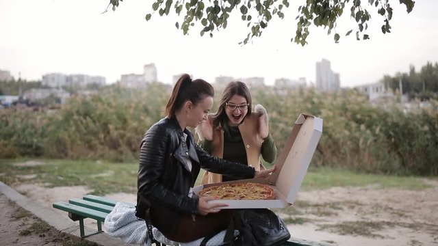 Two women eating pizza in a large box in the city sitting on a bench on the promenade