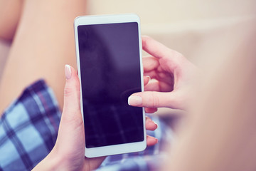 Back view of a woman hands using smart phone with blank screen close up. White phone with a black screen in the hands of a young girl lying on the floor with her feet on the couch